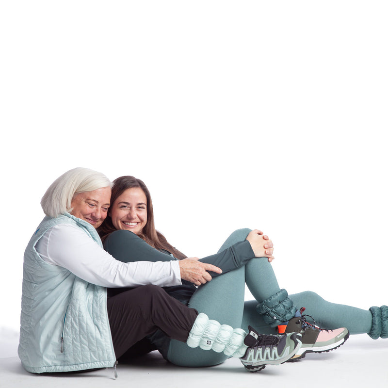 Mother and daughter sitting together with their legs stretches out in front of them. Mom is wearing Alpine Mist Hikas Gaiters with Solomon trail runners, black leggings, a white teeshirt, and a pale aqua vest. Daughter is wearing Salewa hiking boots, green leggings, dark green shirt, and Blue Spruce Hikas Gaiters.