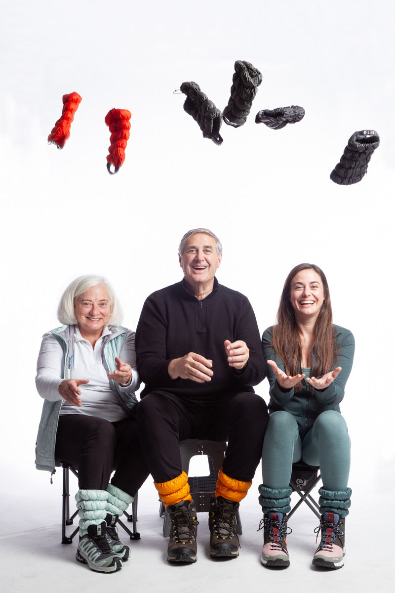 Three people sitting in camp chairs tossing Hikas Gaiters into the air. They're each wearing hiking outfits, plus Hikas Gaiters in Alpine Mist, Golden Hour, and Blue Spruce. The Hikas Gaiters they're tossing in the air are Very Vermillion, Dark Moss, and Black Onyx. 