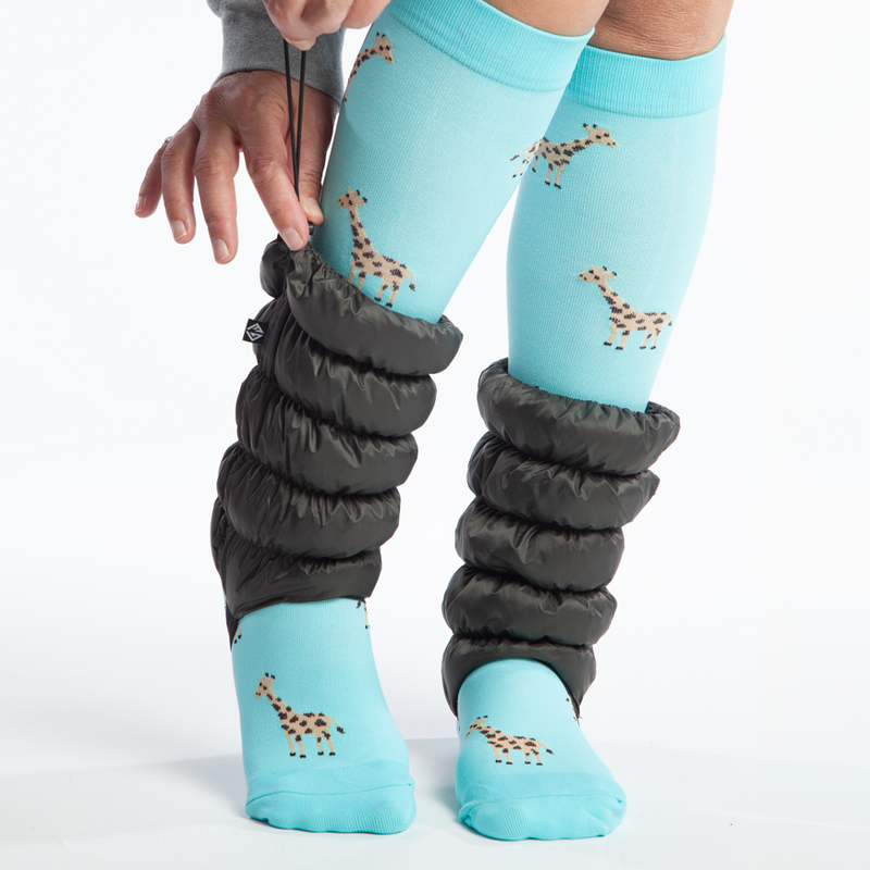 Person in giraffe socks wearing Deep Moss Hikas Gaiters. They have their right heel kicked up and they're pulling the drawstring elastic on the right Hikas Gaiter to tighten the top. 