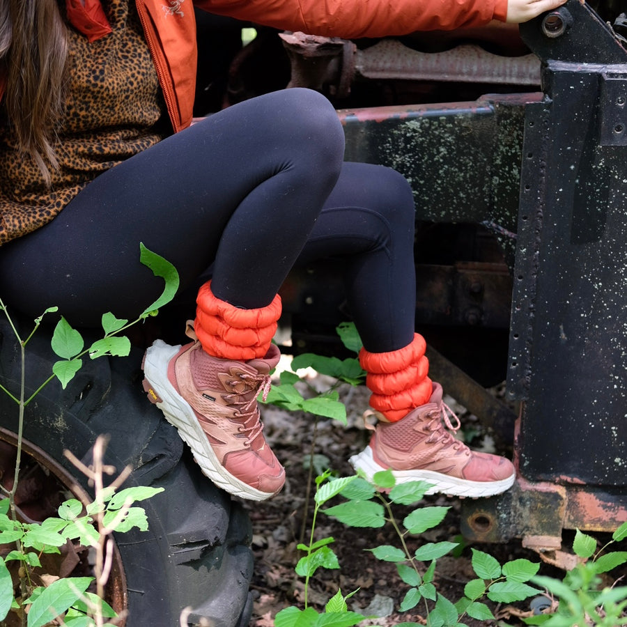 Video cover image, person sitting on a large tractor tire wearing leggings, a leopard print sweater, orange jacket, Very Vermillion Hikas, and pink Hoka hiking boots. 