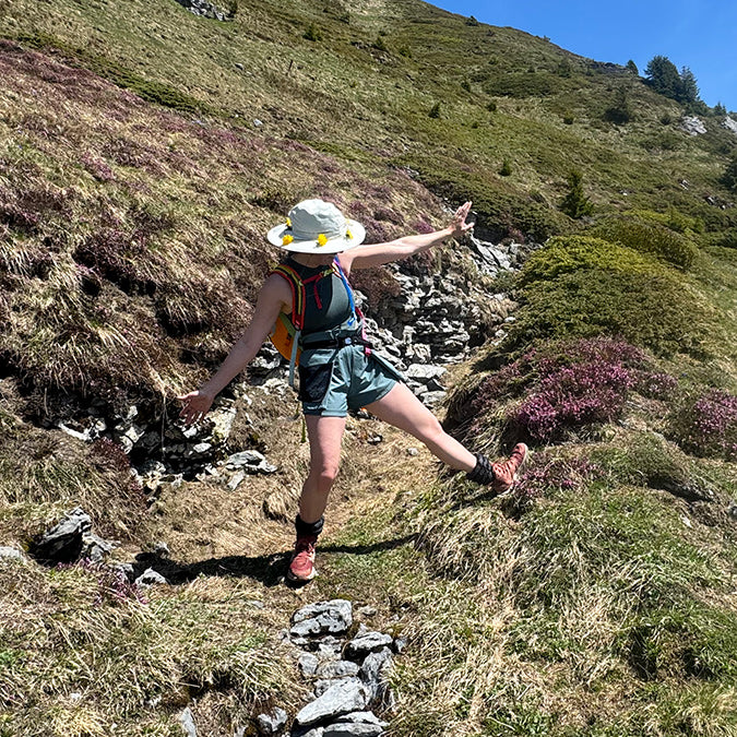 Person strikes a pose mountain side. They have their arms flung wide, and one leg kicking out. They're wearing a large sunhat with a wildflower crown, a daypack, a green tanktop, and green shorts, with Black Onyx Hikas and pink hiking boots.