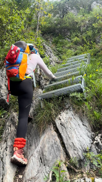 Person wearing backpack and Very Vermillion Hikas Gaiters at the bottom of a ladder, reaching the bottom rung to begin climbing up. 